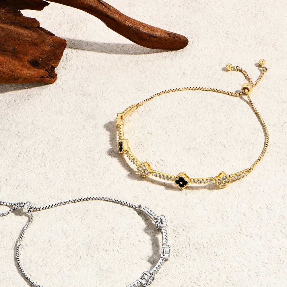 Bee-Bees gold-plated moissanite flower tennis bracelet with adjustable chain and black floral accents on textured sand.