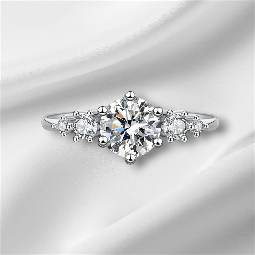 Bee-Bees 1ct round moissanite engagement ring in sterling silver with 18k white gold plating. Features a central solitaire stone flanked by smaller stones.