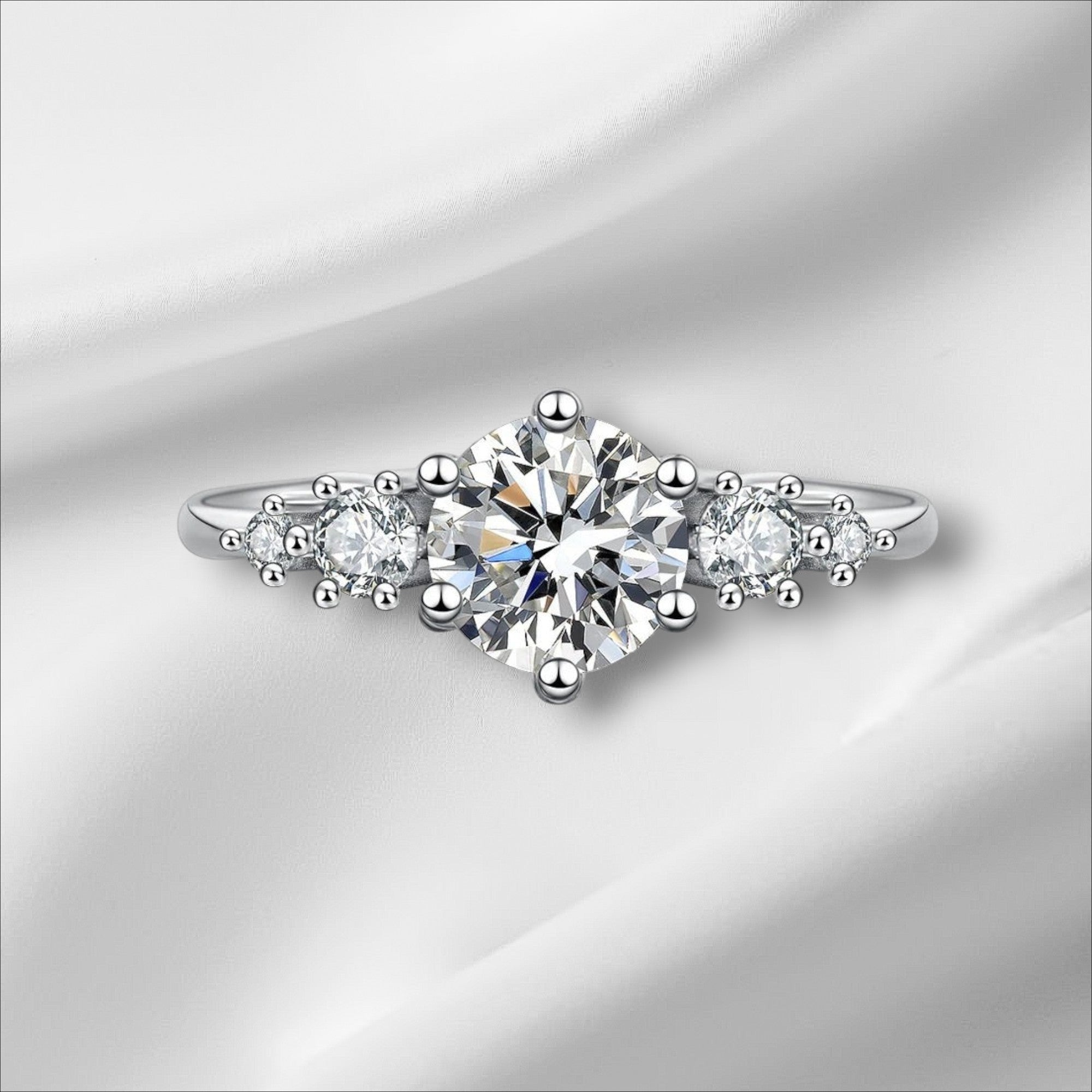 Bee-Bees 1ct round moissanite engagement ring in sterling silver with 18k white gold plating. Features a central solitaire stone flanked by smaller stones.
