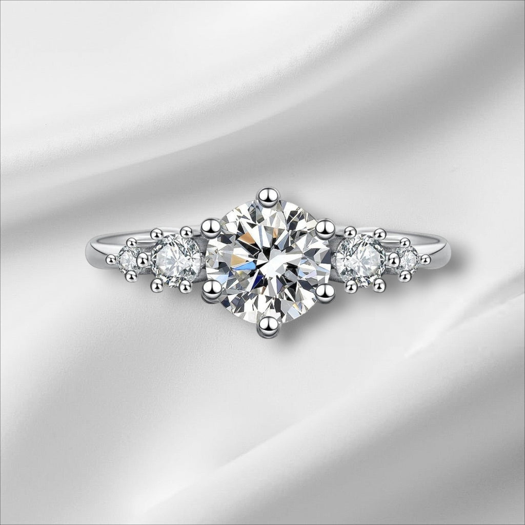 Bee-Bees 1ct round moissanite engagement ring in sterling silver with 18k white gold plating. Features a central solitaire stone flanked by smaller stones.