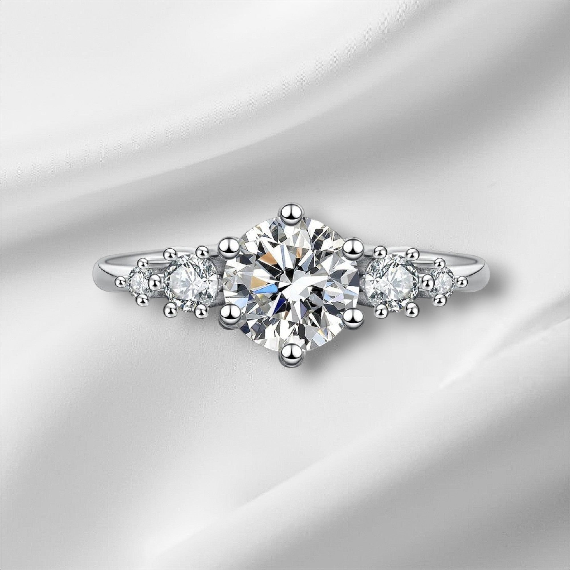 Bee-Bees 1ct round moissanite engagement ring in sterling silver with 18k white gold plating. Features a central solitaire stone flanked by smaller stones.